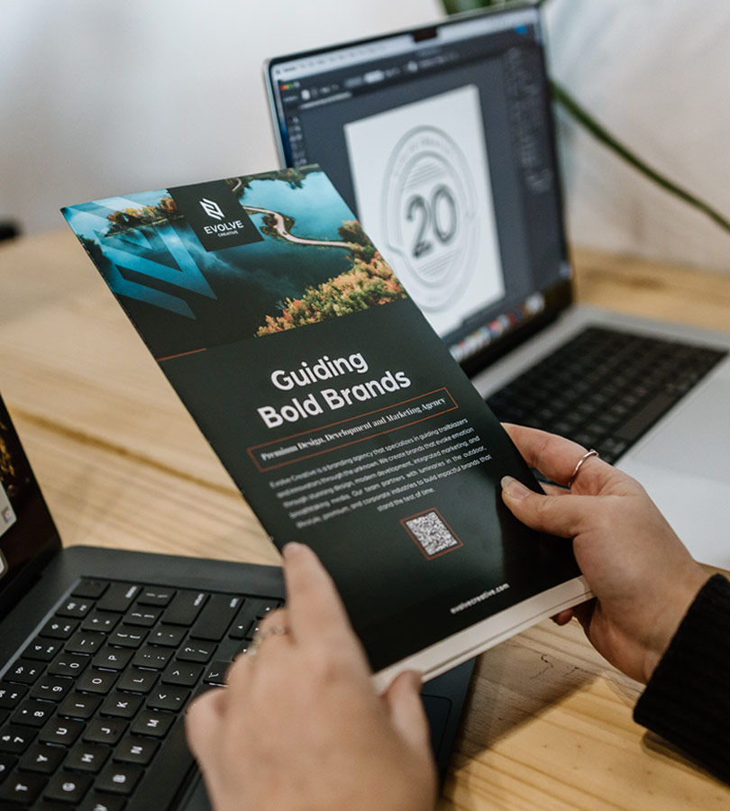 Person holding brochure near laptop on wooden desk.