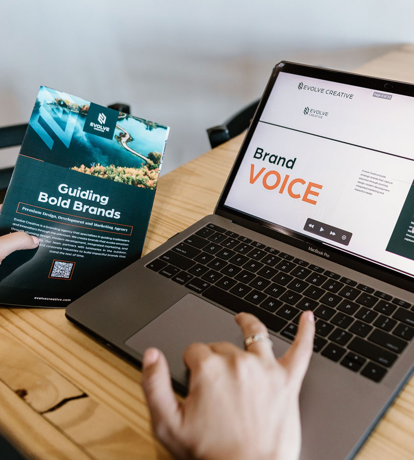 Person holding brand guide, using laptop on marketing strategies.