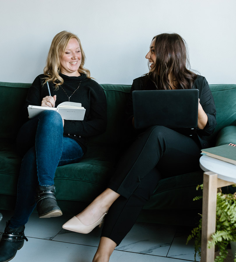 Two women collaborating on couch with laptop and notebook